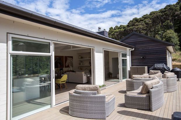 Bright exterior view of a contemporary coastal-style home with a large timber deck, extensive sliding glass doors, white weatherboard walls, and a dark timber-clad outbuilding. The deck is furnished with several wicker lounge chairs and sofas, opening directly into a light-filled living area. Dense mature vegetation surrounds the property.