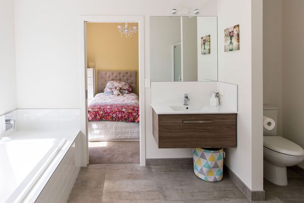Bright modern bathroom with a white bathtub, wall-mounted timber vanity, integrated basin, large mirrored cabinet, toilet, and grey stone-look floor tiles. An adjoining bedroom is clearly visible through the open doorway, featuring a tufted upholstered bedhead, patterned bedding, and a small chandelier.