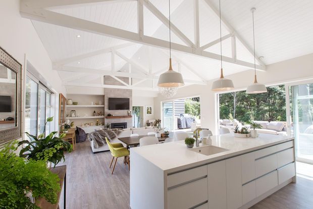 Bright open-plan kitchen and living area with a large white island, integrated sink and cabinetry, pendant lights, exposed white timber roof trusses, and extensive glazing. The adjoining living room includes a sofa, media wall and built-in shelving, creating an airy contemporary coastal-style interior.