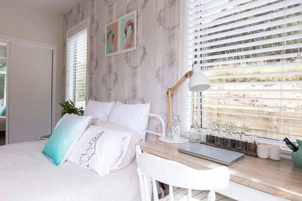 Bright, compact bedroom with a white bed, layered pillows, and a white desk positioned beneath large horizontal blinds. The room features pale floral-patterned wallpaper, white timber-style furniture, a wooden articulated desk lamp, potted plants, and pastel flamingo artwork, creating a fresh coastal-inspired feel.
