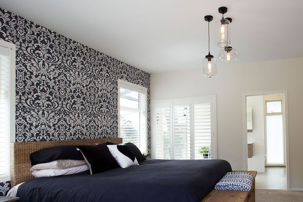 Bright bedroom with a large bed dressed in dark navy and white bedding, a woven rattan headboard, bold black-and-white damask wallpaper on the feature wall, plantation shutters, and three hanging glass pendant lights. A doorway reveals an adjacent bright area with built-in cabinetry.