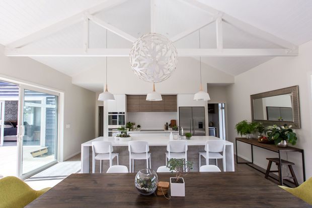 Bright open-plan kitchen and dining area with a vaulted white ceiling, exposed painted timber trusses, a large white island with bar seating, stainless-steel appliances, pendant lighting, and a dark timber dining table. Sliding glazed doors open to an outdoor area, while a console and mirror sit along the right wall.