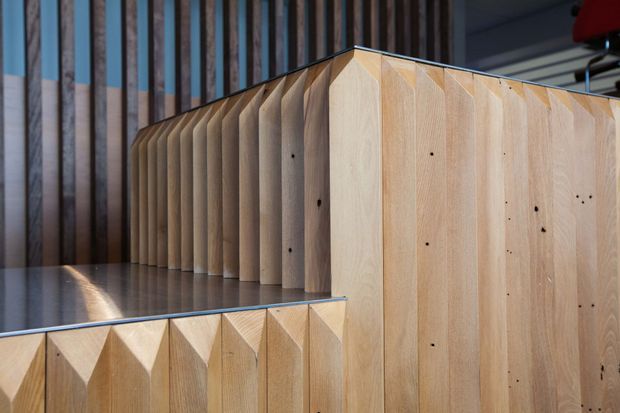 Close-up architectural detail of a sculptural timber staircase or stepped built-in structure. Pale plywood or timber panels form repeated faceted, folded vertical elements along the risers and side enclosure, with visible dark fixing points. A dark grey smooth floor and a backdrop of vertical timber battens add contrast, while cool daylight enters from the left. The composition emphasises geometric joinery, texture, and crafted timber construction.