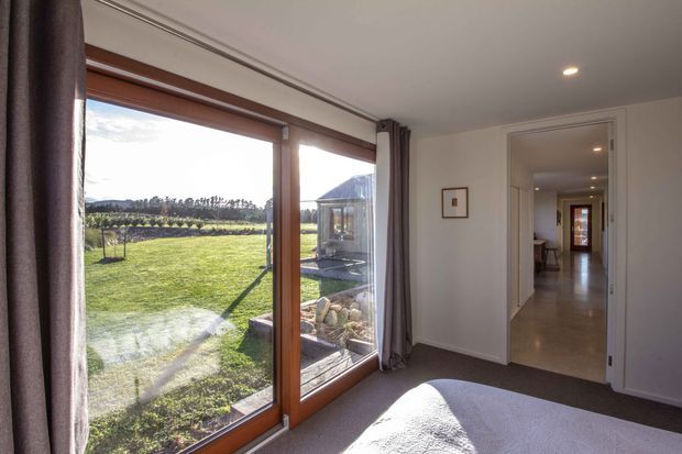 A bright bedroom with large timber-framed glazed doors opening toward a landscaped lawn and garden. The room has neutral painted walls, a dark carpeted floor, recessed ceiling lighting, full-length curtains, and a hallway visible through an open doorway. The exterior garden and a small glazed garden structure are visible beyond the doors.