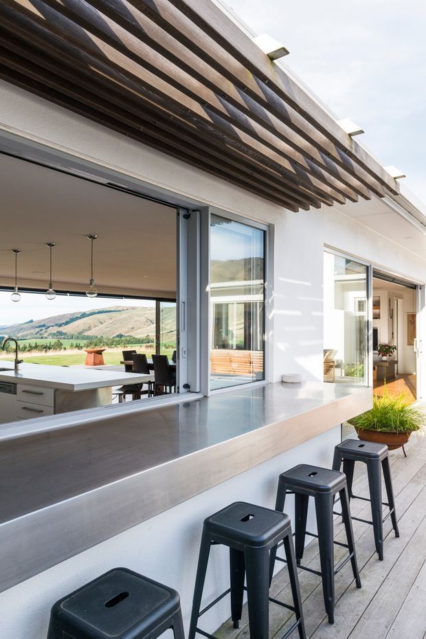 A bright contemporary outdoor entertaining area beside a modern home, featuring a long stainless-steel counter with stools, a pass-through window to the kitchen, timber decking, and a timber pergola. Sliding glass doors open to an interior kitchen and dining area, with rolling hills visible beyond.