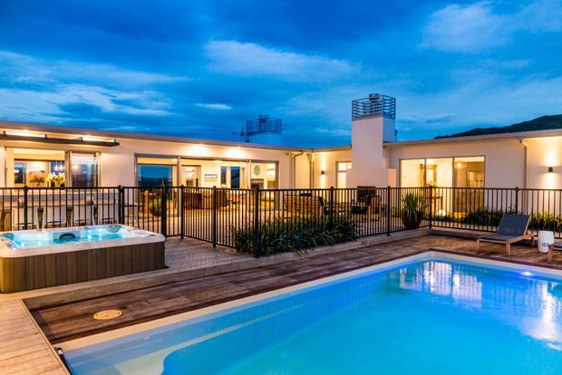 Evening exterior view of a contemporary single-storey residence arranged around a pool terrace. The scene features a bright swimming pool, a raised spa, timber decking, black metal balustrades, warm interior lighting, and a flat-roofed building with a prominent chimney or services volume.
