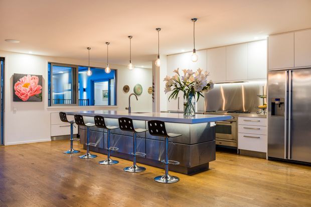 A spacious contemporary kitchen with a large island and five bar stools, integrated stainless-steel appliances, white handleless cabinetry, a reflective metal splashback, timber flooring, and pendant lighting. The open kitchen connects visually to an adjacent hallway or living area, with colourful artwork and a floral arrangement adding decorative accents.