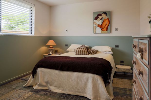 A cozy bedroom with a neatly made bed layered in white bedding, a dark textured throw, and patterned accent cushions. The room has two-tone walls with a muted sage-green lower section and warm off-white upper section, a timber floor partly covered by a patterned rug, a bedside table with a lit lamp, white horizontal window blinds, and a timber chest of drawers. Artwork hangs above the bed.