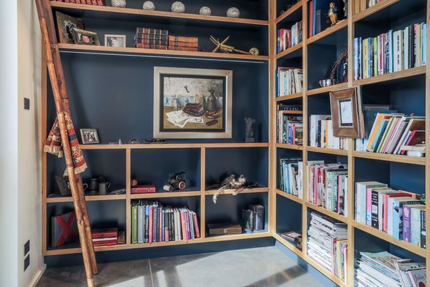 Built-in floor-to-ceiling library shelving wraps around two walls, finished in a deep charcoal-blue with warm timber framing and trim. The shelves display books, framed artwork, ceramics, photographs, and decorative objects, with a timber library ladder leaning against the left side. Natural light falls across the pale hard floor, creating a refined, study-like reading and storage space.