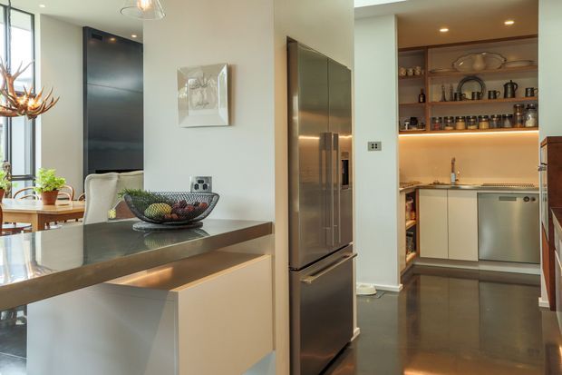 A contemporary kitchen viewed from the main cooking and dining area, with a large stone island in the foreground, integrated stainless-steel refrigerator, pale cabinetry, and a compact secondary kitchen or pantry beyond. The rear space features open timber shelving, under-shelf lighting, a stainless-steel dishwasher, and dark polished flooring. Natural light enters through full-height glazing at left, creating a bright, refined interior with warm timber and metallic accents.