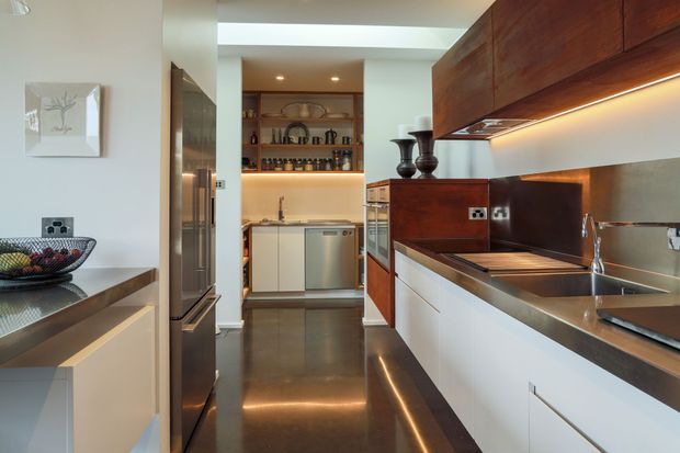 A contemporary galley kitchen with dark polished flooring, white lower cabinetry, rich dark timber overhead cabinetry, stainless-steel appliances and sink, and warm integrated under-cabinet lighting. A compact pantry or scullery is visible at the rear, with open shelving and a skylight contributing to the bright, functional space.