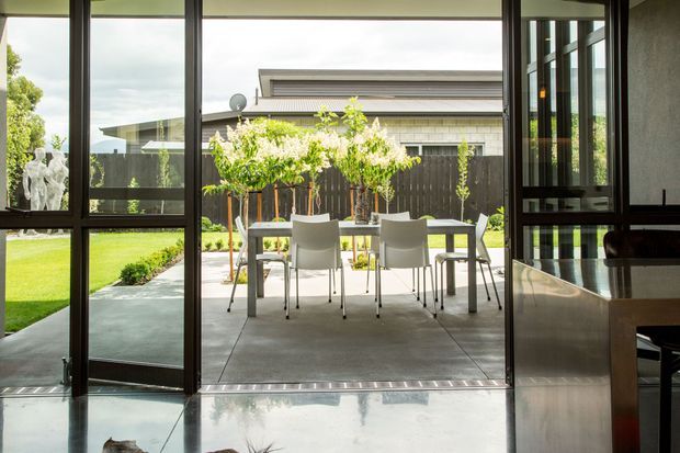 A contemporary indoor-outdoor dining area viewed from inside through fully open dark-framed glazed doors. A simple dining table and chairs sit on a paved terrace, opening onto a manicured lawn and planted garden. The interior foreground includes polished concrete flooring and a section of sleek built-in cabinetry, while the exterior shows a dark timber fence and a neighbouring contemporary building.