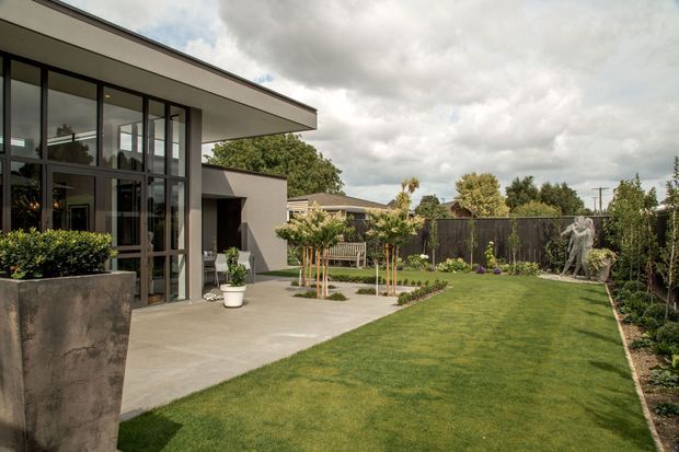 Contemporary residential rear courtyard with a flat-roofed extension, extensive floor-to-ceiling dark-framed glazing, a concrete terrace, manicured lawn, raised planting beds, mature garden borders, and a dark timber privacy fence. The outdoor space is viewed from the garden toward the house under an overcast sky.