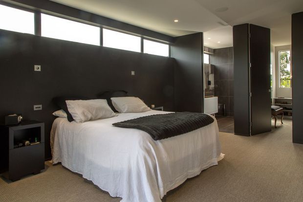 A contemporary bedroom with a neatly made double bed, white bedding, dark charcoal throw, and dark built-in wall panels. High clerestory windows bring in natural light, while a carpeted floor and minimal black bedside storage create a calm, restrained interior. A partially visible bathroom and adjoining living area appear beyond the open partition.