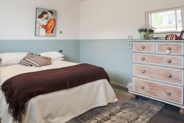 A cozy bedroom with a white bed dressed in a dark brown throw and patterned accent pillow, set against a two-tone pale blue and white wall. A distressed painted timber chest of drawers, indoor plants, artwork, and a patterned area rug add character to the space. Natural light enters through a horizontal window with blinds.