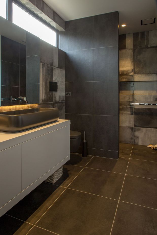 A contemporary dark-toned bathroom with large charcoal and textured metallic-look wall tiles, matching floor tiles, a floating light-grey vanity with a dark vessel basin, wall-mounted tapware, a large mirror, toilet, and walk-in shower. Warm concealed lighting beneath the vanity and in the shower niche contrasts with the moody finishes. A high horizontal window provides natural light.