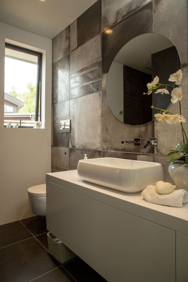 A contemporary bathroom featuring a wall-mounted grey vanity with a white vessel basin, wall-mounted chrome tapware, a round mirror, toilet, dark floor tiles, and a dramatic patchwork metallic grey tiled feature wall. A window provides natural light, while an orchid adds a small decorative accent.
