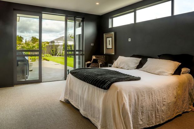 A contemporary bedroom with a large bed dressed in white linen and a dark textured throw, set against charcoal walls. Full-height glazed doors open to an outdoor patio and garden, while high clerestory windows bring in natural light. Neutral carpet, a framed artwork, and a small occasional chair complete the restrained interior.