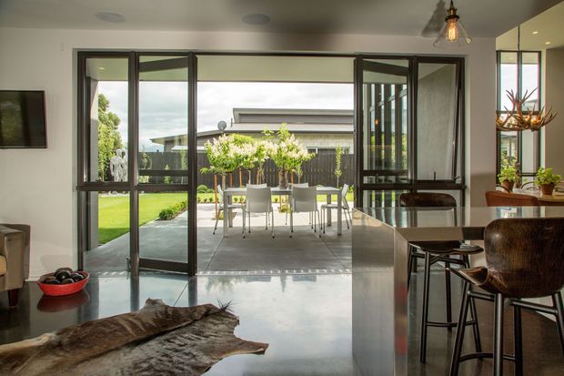 A contemporary open-plan interior with a polished grey concrete floor, dark-framed full-height glazed doors opening onto a paved patio and lawn, and a kitchen island with dark bar stools in the foreground. The room includes a dining setting outdoors, warm timber and leather-toned furnishings, indoor plants, and an exposed pendant light. The strong indoor-outdoor connection and clean-lined glazing dominate the view.