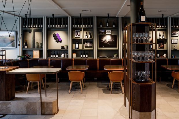 A refined wine bar or restaurant interior with a long upholstered banquette, timber dining tables and chairs, a marble-look tiled floor, and extensive backlit shelving displaying wine bottles and glassware. Dark timber joinery, framed artwork, pendant lighting, and a white paneled ceiling create an intimate, sophisticated atmosphere.