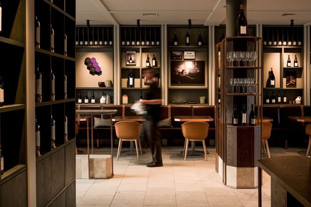 A sophisticated wine bar or tasting room with extensive built-in bottle displays, dark timber cabinetry, illuminated shelving, framed artwork, and a central freestanding wine display rack. A bar-height counter with stools and upholstered seating occupies the room, while warm ambient lighting and pale tiled flooring create an upscale hospitality atmosphere.
