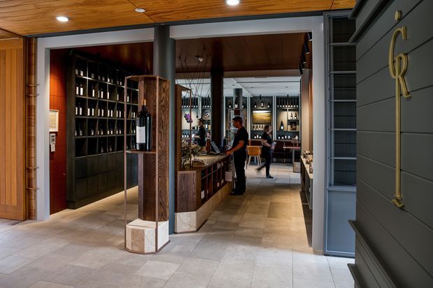 A refined wine shop or tasting-room interior viewed from the entrance, with extensive dark bottle display shelving, a central timber-clad service counter, display pedestals, warm timber ceilings, stone-look flooring, and dark grey architectural joinery. Staff and customers are visible deeper in the space.