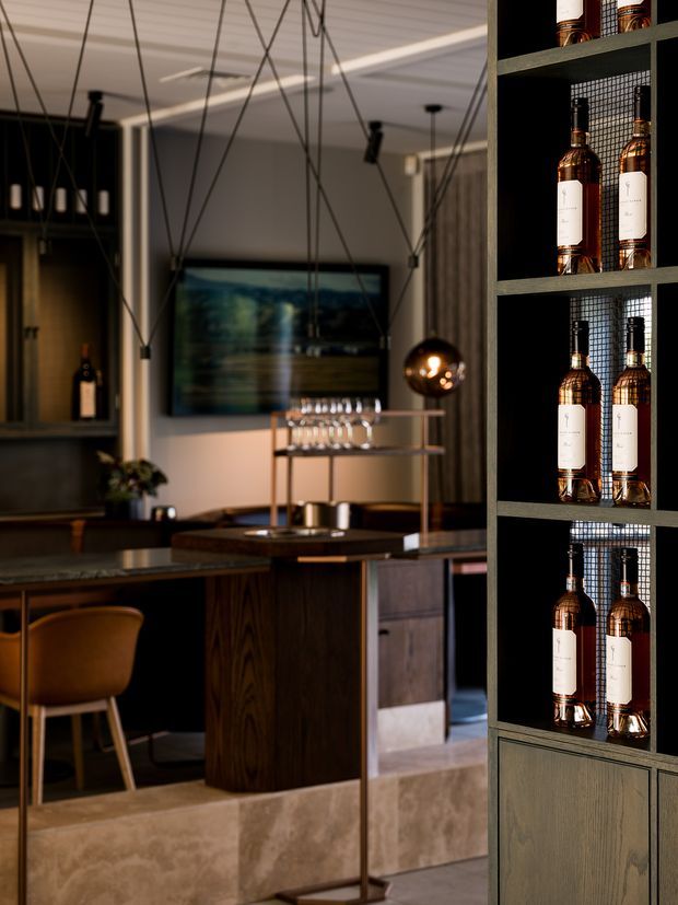 A sophisticated home bar with dark timber cabinetry, a central island, glassware shelving, hanging pendant lights, and a prominent built-in display of liquor bottles. Warm ambient lighting, bronze and timber finishes, and a large artwork create a moody, refined atmosphere.