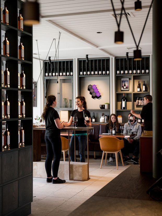 A stylish wine bar or tasting room viewed from the entrance, with dark built-in bottle shelving, a central standing-height counter, booth seating, mustard upholstered chairs, and pendant lighting. The space features a light tiled floor, dark timber cabinetry, a white panelled ceiling, and a warm, contemporary hospitality atmosphere. People are socialising and staff are serving wine.