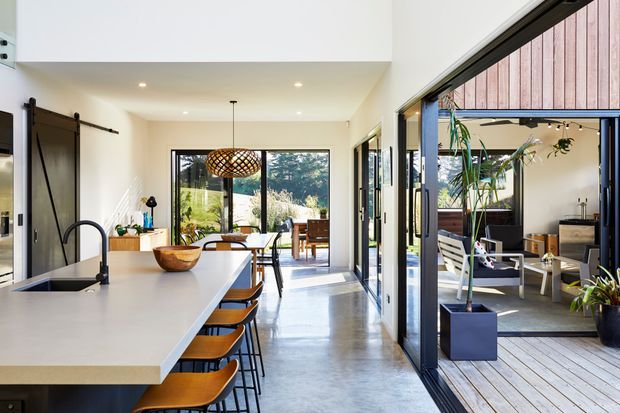 Bright, open-plan contemporary kitchen and dining area with a long pale island, integrated sink and black tap, timber bar stools, polished concrete flooring, and expansive black-framed glazing. The glazing opens onto a timber deck and covered outdoor lounge, with landscaped greenery visible beyond. A sculptural woven pendant hangs over the dining table, and a barn-style sliding door adds a strong architectural feature.
