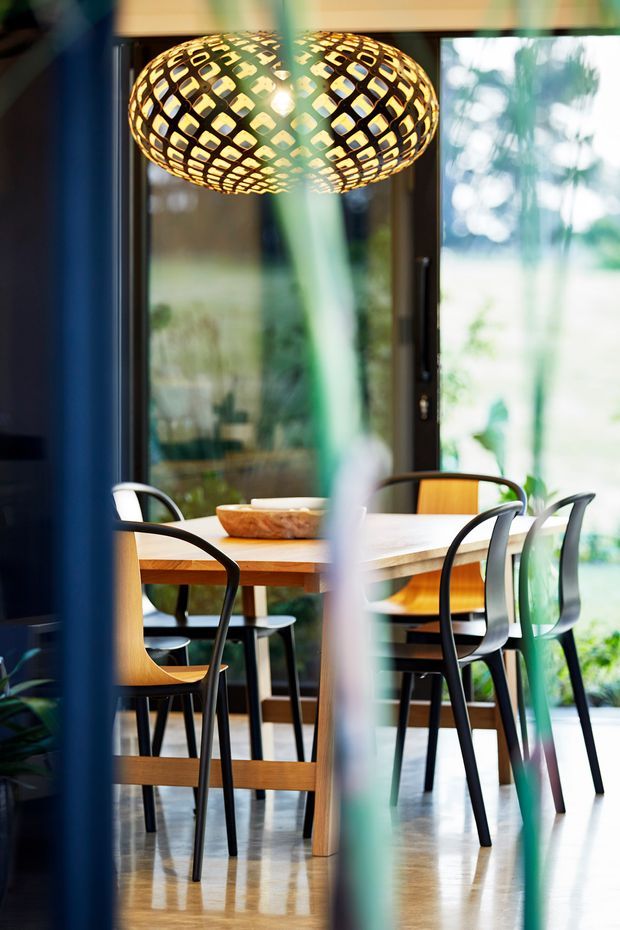 A dining area viewed through partially obscuring foreground elements, featuring a rectangular timber dining table, mixed black and timber dining chairs, polished light-toned flooring, large glazed doors or windows, and a prominent woven oval pendant light. The space has a warm, contemporary feel with strong indoor–outdoor connection and greenery visible beyond the glazing.
