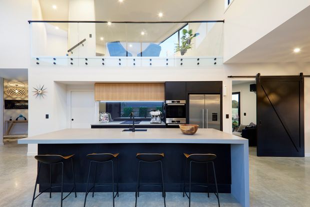 Bright, open-plan contemporary kitchen with a large island and four stools, integrated dark cabinetry, stainless-steel appliances, pale stone or composite benchtops, and a double-height void. A glazed mezzanine balustrade and stair are visible above, with large openings and polished concrete flooring contributing to the spacious modern interior.