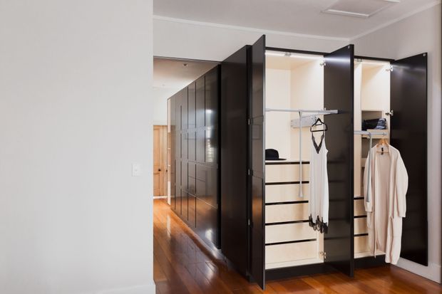 A contemporary walk-in wardrobe with tall glossy dark cabinetry, open hanging rails, drawers, shelving, and integrated lighting. The wardrobe opens onto a polished timber-floored hallway with a timber door visible in the background. The space is bright, orderly, and refined.