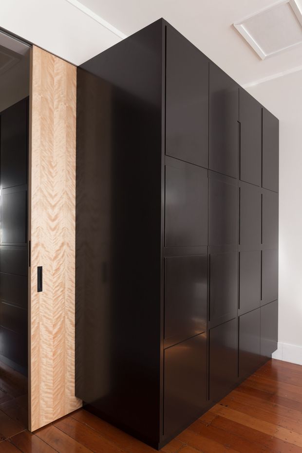 A tall, built-in dark cabinetry wall or wardrobe occupies the corner of a bright interior. The cabinetry has flat-panel matte dark fronts with subtle geometric divisions and integrated handles, paired with a contrasting light timber veneer door featuring a chevron grain. It stands on a warm timber floor beside white walls and a white ceiling with simple trim and a ceiling access panel.