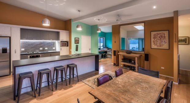 Open-plan kitchen and dining area with a long island breakfast bar, dark cabinetry, stainless-steel appliances, mosaic tile splashback, timber flooring, and industrial-style stools. The adjoining dining zone features a large rustic timber table and leather dining chairs, while a living area and bright blue-accented room are visible beyond.
