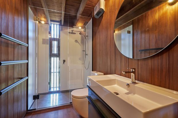 A compact, richly detailed bathroom with dark timber wall panelling and ceiling, a large white double vanity, round mirror, toilet, and a fully glazed walk-in shower. The shower includes a rain shower, handheld fitting, and narrow vertical window, while warm integrated lighting creates a refined, timber-lined interior.
