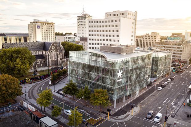 Aerial view of a contemporary multi-storey glass commercial building at a city intersection, with a curved glazed corner, rooftop terrace, and prominent white star logo. Surrounding urban streets, mature trees, mid-rise office buildings, and a historic Gothic church are visible.