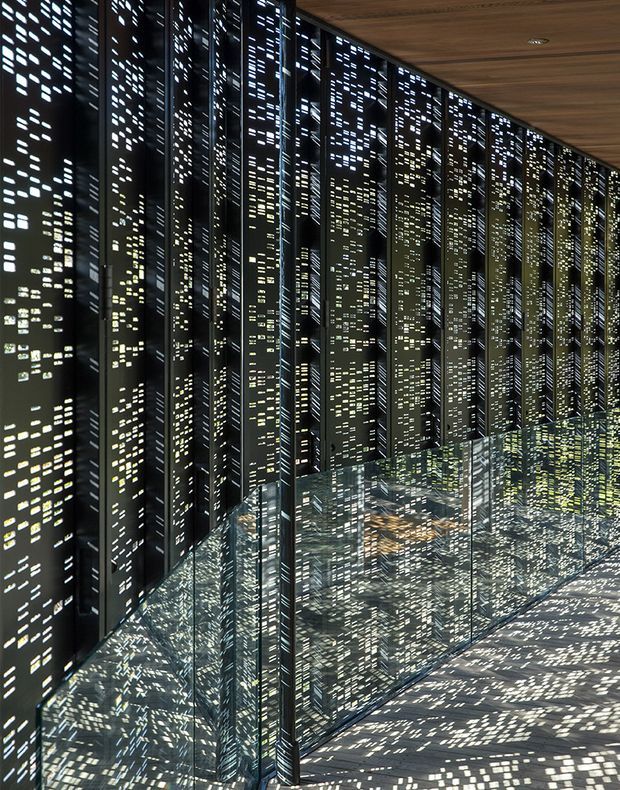 A dramatic architectural interior featuring a long wall of dark vertical perforated screens or fins casting dense, pixel-like patterns of light across the glossy floor and glass balustrade. The space has a dark, reflective, highly geometric appearance with a warm timber ceiling and a contemporary installation-like quality.