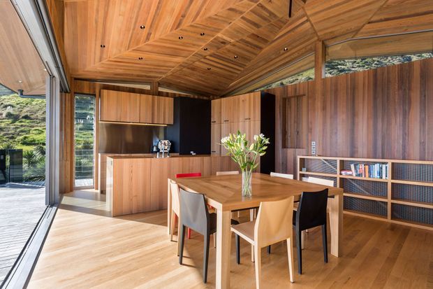 A warm, timber-lined open-plan dining and kitchen interior with extensive floor-to-ceiling glazing opening toward a deck and green landscape. The room features a central timber dining table with mixed chairs, a compact timber kitchen with island, integrated cabinetry, a built-in open shelving unit, and a sculptural angled timber ceiling with recessed spotlights.