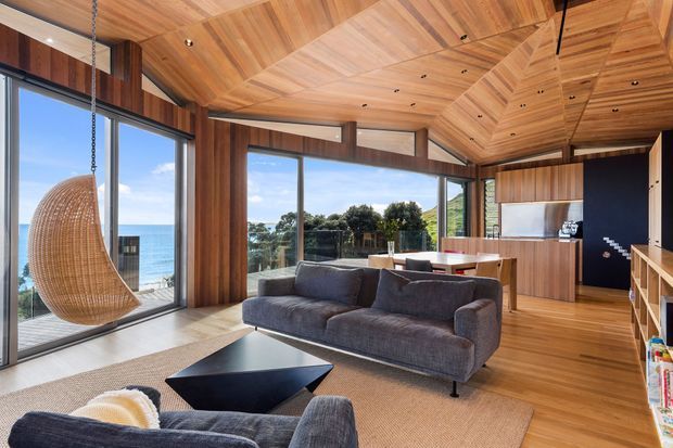 A warm, contemporary open-plan living area with extensive ocean-facing glazing, a timber-lined angular ceiling, timber wall panelling and flooring, and a dark charcoal sectional sofa arranged around a geometric black coffee table. A suspended woven egg chair sits beside the glazing, while a dining area and compact kitchen are visible beyond.