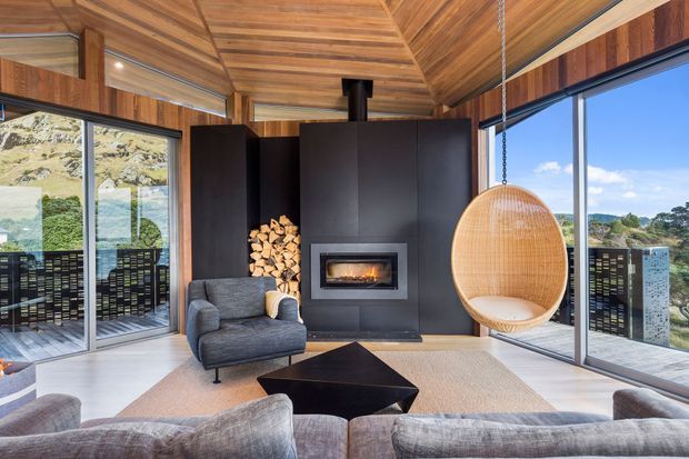 A contemporary living room with expansive floor-to-ceiling glazing, a dramatic black fireplace wall with integrated firebox and stacked firewood, warm timber walls and a vaulted timber ceiling. The room includes a grey upholstered sofa and armchair, a low black coffee table, a woven rug, and a suspended woven egg chair. Mountainous landscape is visible beyond the glazing and balcony.