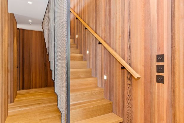 A narrow modern stairway and hallway clad extensively in warm vertical timber. The staircase has matching timber treads and a simple timber handrail, with a dark-framed perforated metal or mesh balustrade. Recessed step and ceiling lighting, timber wall panelling, and dark electrical switch plates create a clean contemporary interior.