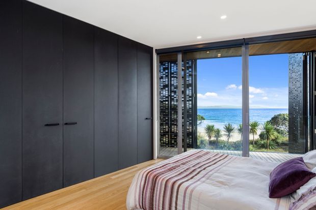 Contemporary bedroom with a wall of dark built-in wardrobes, pale timber flooring, and a large glazed opening overlooking a bright coastal landscape. The bed has striped bedding and a deep purple accent cushion, while perforated privacy screens frame the view.