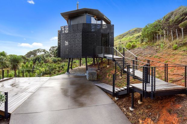 Exterior view of a compact elevated modern building set into a steep landscaped hillside. The structure features dark perforated metal screening, slim steel supports, a shallow projecting roof, and an external staircase and walkway leading to a broad concrete terrace. The surrounding setting includes dense vegetation, exposed reddish earth, and expansive blue sky.