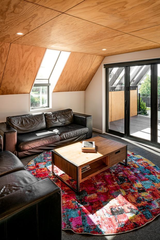 A compact, sunlit living room with a distinctive plywood-lined sloped ceiling and angled wall panels. The room contains dark brown leather sofas and ottomans, a rectangular timber coffee table, and a highly colourful patterned area rug. Black-framed full-height sliding glass doors open to a timber deck, while a narrow skylight and window bring in strong natural light.