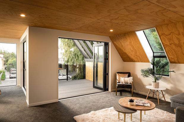 A warm, contemporary living room or lounge with a dramatic sloping plywood-lined ceiling and matching timber wall surfaces. The room has grey carpet, a neutral painted wall, recessed downlights, large black-framed glazing and a glazed door opening onto a timber deck or courtyard. A compact seating arrangement, round coffee tables, rug, armchair and potted plant create a relaxed, inviting interior. A roof window brings in additional daylight.