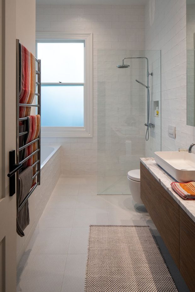 Bright, narrow contemporary bathroom with full-height pale tiled walls and matching tiled floor. The room includes a walk-in glass shower with chrome fixtures, a freestanding or partially visible bathtub beneath a frosted window, a wall-mounted heated towel rail with striped towels, a floating timber vanity with a pale stone benchtop and inset basin, and a toilet. The palette is light and neutral with warm timber and colourful textile accents.