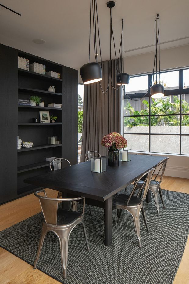 A contemporary dining room with a large dark timber dining table surrounded by industrial-style metal chairs. A full-height built-in dark shelving unit displays books and decorative objects, while multiple black pendant lights hang at varied heights above the table. Large black-framed windows, full-length curtains, light timber flooring, and a textured charcoal rug complete the room.