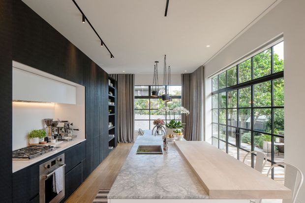 A bright, elongated contemporary kitchen with a large stone island and adjoining timber breakfast bar. Dark vertical timber cabinetry lines one wall, incorporating open shelving, a concealed rangehood and integrated cooking appliances. Full-height black-framed glazing opens the room visually to greenery, while pendant lights, track lighting and warm timber flooring complete the refined fit-out.