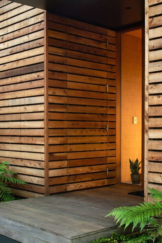 A close view of a contemporary timber-clad entryway with a raised grey-stained timber deck and steps. The entrance is framed by horizontal weathered wood boards, with a large slatted timber pivot door, warm timber-lined interior, dark ceiling, recessed lighting, and a potted plant. Ferns and low greenery surround the deck, creating a natural, sheltered arrival space.