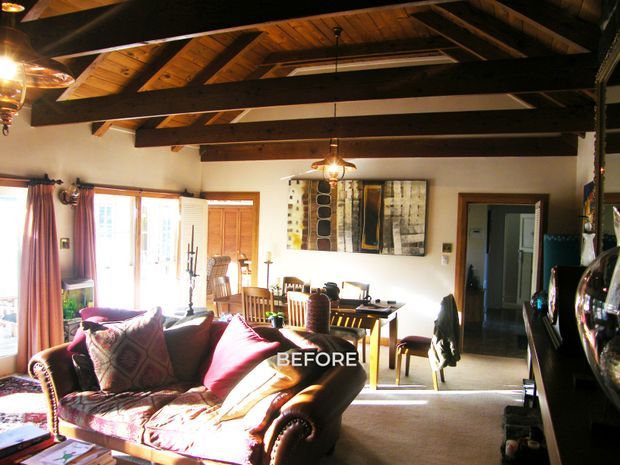 A warmly lit open-plan living and dining area photographed from the living room. The room features a patterned traditional sofa, timber dining table and chairs, exposed dark timber roof beams with a pitched ceiling, timber-framed glazed doors, carpeted flooring, and a large abstract wall artwork. A hallway or doorway is visible at the rear, and the image is labelled “BEFORE.”
